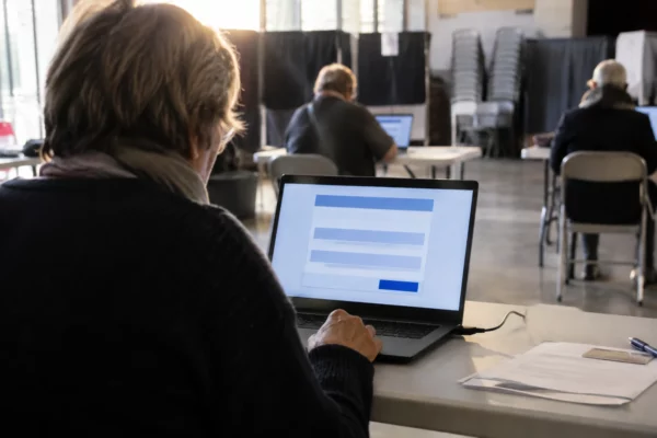 Un ordinateur portable affiche une interface de vote générique, installé dans une salle avec d’autres postes et des chaises empilées.